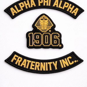 Może przedstawiać: Czarne i złote haftowane naszywki. Górna naszywka zawiera napis "ALPHA PHI ALPHA" w łukowatym kształcie. Poniżej znajduje się naszywka ze złotym emblematem kobry i numerem "1906". Dolna naszywka zawiera napis "FRATERNITY INC." w kolorze złotym.