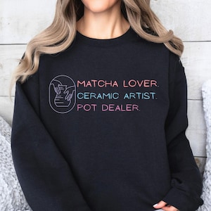 May include: A black crewneck sweatshirt featuring a graphic on the chest. The graphic includes a circular line drawing of hands shaping a pot, followed by the text: MATCHA LOVER. CERAMIC ARTIST. POT DEALER. The text is in pink and light blue.