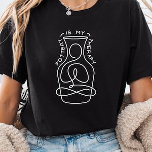 May include: A black short-sleeved t-shirt featuring a white line art graphic of a person in a seated meditation pose inside a vase shape. The text "POTTERY IS MY THERAPY" is printed in white, arching around the top of the vase illustration.
