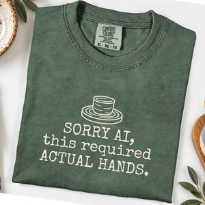 May include: A forest green crewneck t-shirt featuring a white line-art graphic of a pottery wheel and the text "SORRY AI, this required ACTUAL HANDS." The shirt has a Comfort Colors brand label at the inner neckline indicating size XL.
