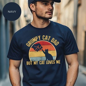 May include: Navy blue t-shirt with the text "GRUMPY CAT DAD" above a graphic of a cat reaching towards a hand, set against a sunset design. The text "BUT MY CAT LOVES ME" is below the graphic.