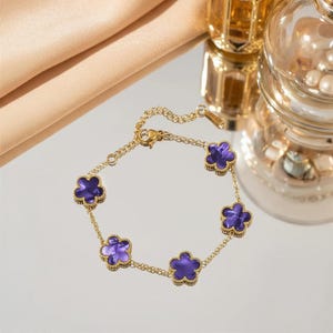 Puede incluir: Una pulsera dorada con seis colgantes morados en forma de flor. Los colgantes están delineados en dorado y conectados por una delicada cadena dorada. La pulsera se muestra sobre una superficie reflectante.