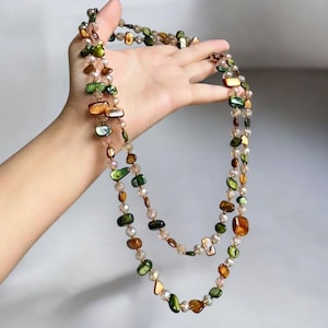 Puede incluir: Un collar largo de varias capas con una mezcla de cuentas coloridas de forma irregular en tonos verdes, marrones y tonos iridiscentes. El collar también incluye cuentas más pequeñas, claras y facetadas, creando un accesorio delicado y elegante.
