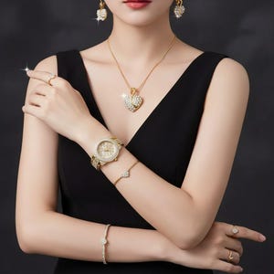 Puede incluir: Un conjunto de joyas doradas con un collar con colgante en forma de corazón, pendientes a juego, un reloj de pulsera, anillos y pulseras. Las joyas están adornadas con cristales brillantes, creando un look glamuroso. La mujer lleva un vestido negro.