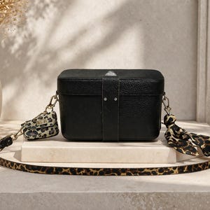 Pode incluir: Uma mala tiracolo retangular preta com textura, uma tira vertical central e ferragens prateadas. A mala apresenta uma alça comprida com padrão de leopardo, uma pequena bolsa a condizer presa na lateral e um lenço com padrão de leopardo.