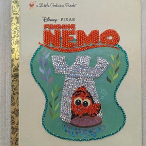 May include: A Little Golden Book titled "Finding Nemo" with a Disney Pixar logo. The cover features a bedazzled Nemo design in a fishbowl, with the title in orange rhinestones. The book has a gold spine with illustrations.
