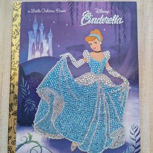 May include: A Little Golden Book titled "Disney Cinderella." The cover features Cinderella in a sparkling blue gown, with a castle in the background. The title is embellished with rhinestones. The book has a gold spine.
