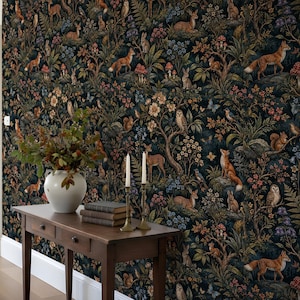 May include: A dark wallpaper featuring a repeating forest pattern with foxes, deer, rabbits, owls, squirrels, and mushrooms. In front, a wooden console table holds a white ceramic vase with green branches and two tall, thin, cream-coloured candles.