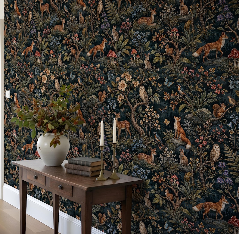May include: A dark wallpaper featuring a repeating forest pattern with foxes, deer, rabbits, owls, squirrels, and mushrooms. In front, a wooden console table holds a white ceramic vase with green branches and two tall, thin, cream-coloured candles.