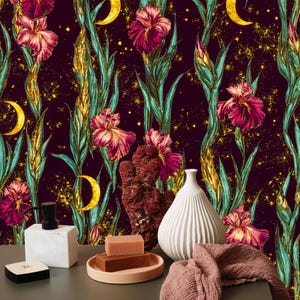 May include: A decorative arrangement featuring a floral wallpaper backdrop with irises, moons, and gold accents. In the foreground, a white vase, soap, and a pink towel are displayed on a gray surface. A black soap dispenser and a small black box are also present.