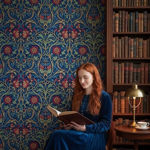 May include: A long-sleeved blue velvet dress worn by a person with long red hair reading an open book. The background features dark blue wallpaper with a repeating floral pattern of red flowers and green leaves, alongside a wooden bookshelf.