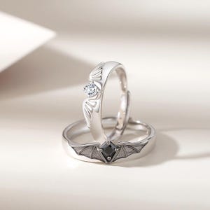 May include: Two silver rings, one with a clear gemstone and wing-like design, the other with a black square gemstone and bat wing design. The rings are set against a soft, neutral background.