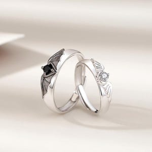 May include: Two silver rings with bat wing designs. One ring features a black square gemstone, the other a clear round gemstone. The rings are set against a soft, neutral background.