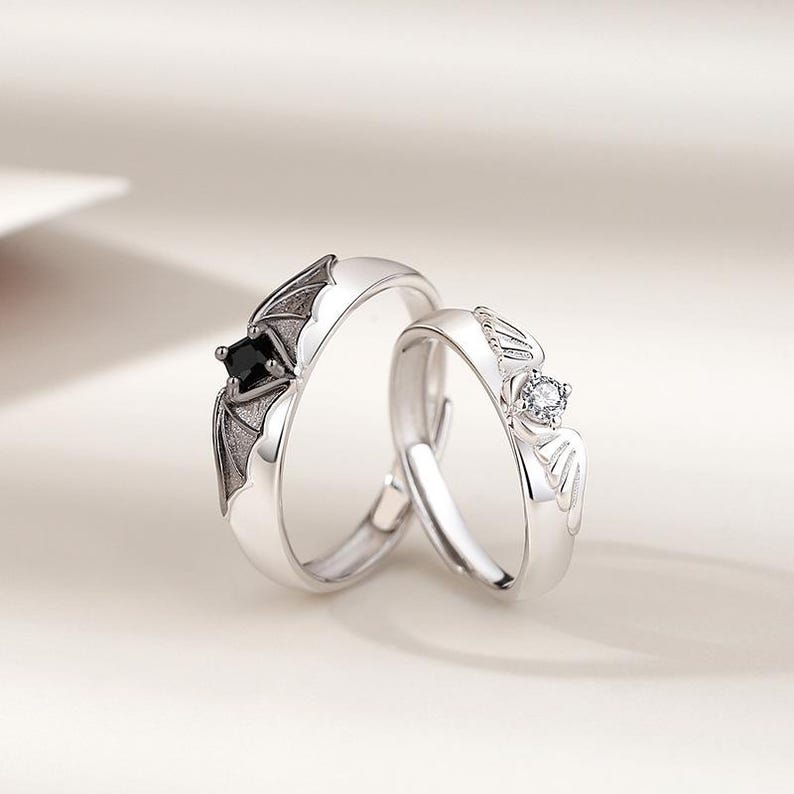 May include: Two silver rings with bat wing designs. One ring features a black square gemstone, the other a clear round gemstone. The rings are set against a soft, neutral background.