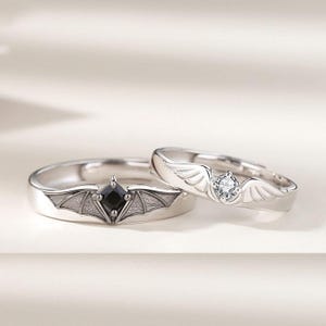 May include: Two silver rings. One ring features a black square gemstone set within a bat wing design. The other ring has a clear round gemstone and wing accents. Both rings are on a white surface.