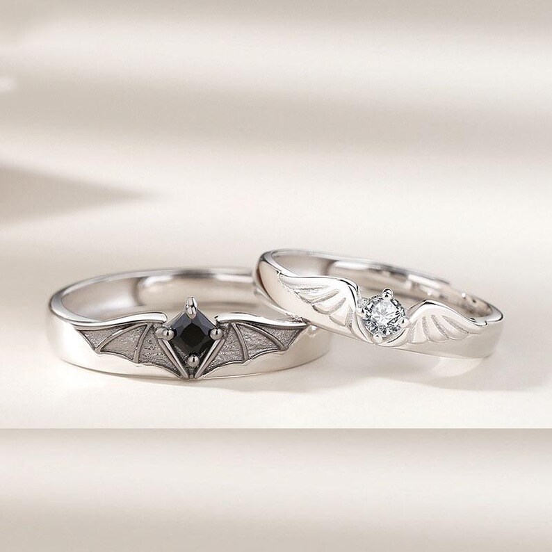 May include: Two silver rings. One ring features a black square gemstone set within a bat wing design. The other ring has a clear round gemstone and wing accents. Both rings are on a white surface.