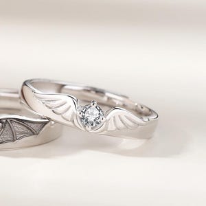 May include: Two silver rings, one with a wing design and a central clear gemstone, the other with a bat wing design. The rings are set against a soft, neutral background, highlighting the intricate details of the jewelry.
