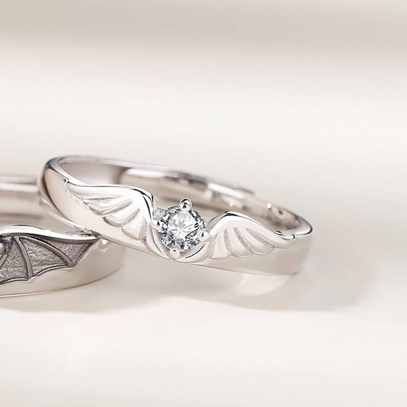 May include: Two silver rings, one with a wing design and a central clear gemstone, the other with a bat wing design. The rings are set against a soft, neutral background, highlighting the intricate details of the jewelry.