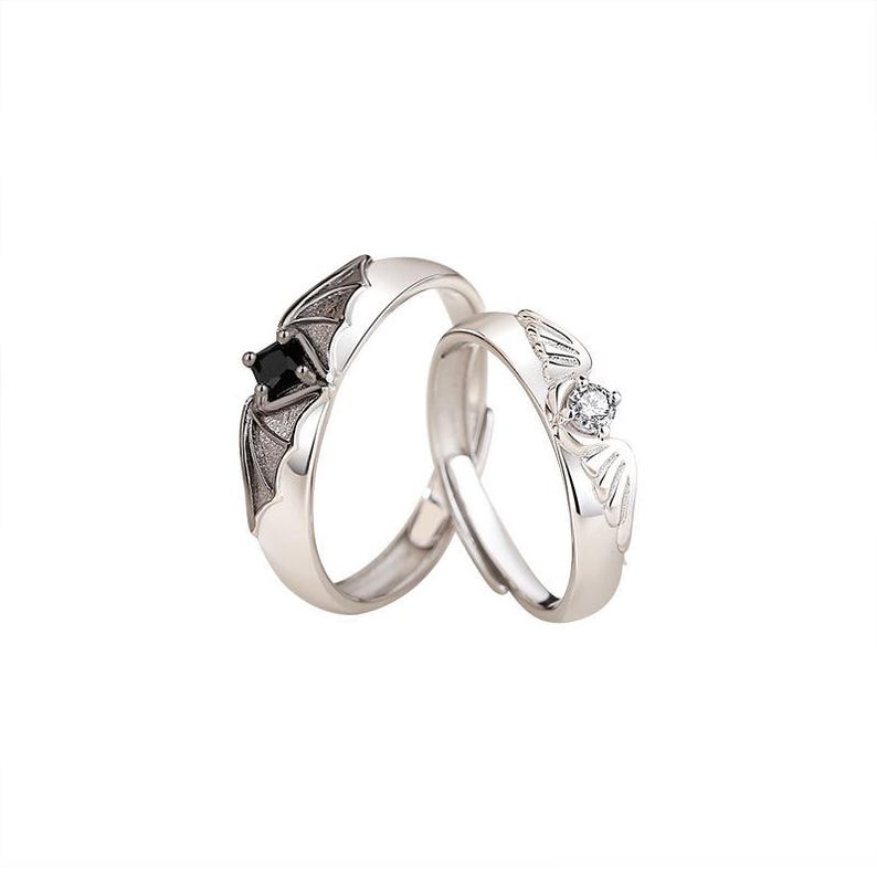 May include: Two silver rings, one with a bat wing design and a black square stone, the other with a wing design and a clear round stone. The rings are set against a white background.