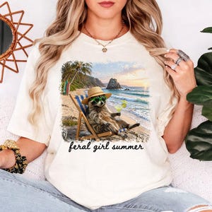 May include: Cream-colored t-shirt featuring a graphic of a raccoon in sunglasses and a straw hat, relaxing on a beach chair with a cocktail. The graphic includes the text "feral girl summer".