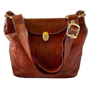 May include: A cognac-colored leather shoulder bag featuring a central embossed logo, a gold-tone metal clasp closure, and a wide adjustable strap with a large gold-tone buckle. The bag has visible stitching details and metallic hardware accents.