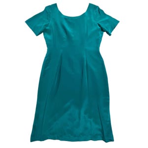 May include: A solid teal-colored dress featuring a scoop neckline, short sleeves, and a knee-length hemline. The garment is constructed with a smooth, slightly reflective fabric that creates subtle vertical draping and a structured silhouette.