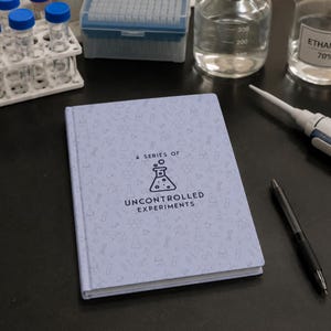 May include: A light blue hardcover notebook featuring a repeating pattern of science-themed icons and the text A SERIES OF UNCONTROLLED EXPERIMENTS centered on the front cover, accompanied by a black pen resting on the surface next to the book.