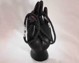 May include: A black beaded necklace with silver-tone clasps is draped over a black hand-shaped display. The necklace is made of small, dark beads, creating a textured appearance. The background is a soft, off-white color.