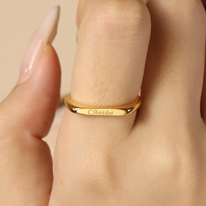 Peut inclure: Une bague dorée polie dotée d'un anneau lisse et légèrement incurvé, avec le prénom Charles gravé en écriture cursive sur la surface avant. Le métal présente une finition réfléchissante et un design simple et épuré entourant le doigt.