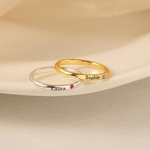 May include: Two thin metal rings, one silver-toned and one gold-toned. The silver ring is engraved with the name Laura and features a small pink stone. The gold ring is engraved with the name Sophie and features a small light blue stone.