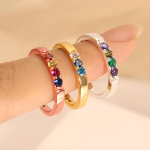 May include: Three metal rings in rose gold, yellow gold, and silver tones, each featuring a row of three small, round, multi-colored gemstones. The bands are smooth and polished, showcasing vibrant red, blue, green, and clear stones.