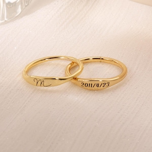 Puede incluir: Dos anillos dorados entrelazados. Uno lleva la letra "m" grabada, el otro la fecha "2011/4/23". El fondo claro resalta los anillos, sugiriendo una joya personalizada y elegante, ideal para ocasiones especiales.