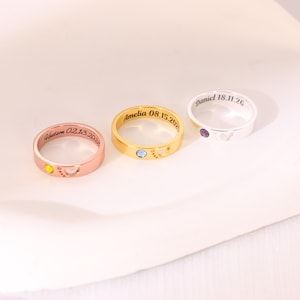 Puede incluir: Tres anillos de metal en tonos oro rosa, oro amarillo y plata. Cada anillo presenta un nombre y fecha grabados, una pequeña piedra preciosa circular y un recorte en forma de corazón. Los grabados dicen Hudson 02.13.2025, Amelia 08.15.2025 y Daniel 18.11.26.