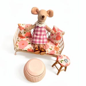 May include: A plush mouse doll wearing a pink checkered dress and sandals sits on a miniature sofa with floral cushions. A matching stool and a pink, textured table complete the scene, all set against a white background.