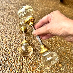 Può includere: Due pomelli per porta in vetro trasparente con taglio sfaccettato simile a un diamante. Ogni pomello presenta una base circolare in metallo color ottone e un lungo e sottile perno metallico che collega le due estremità.