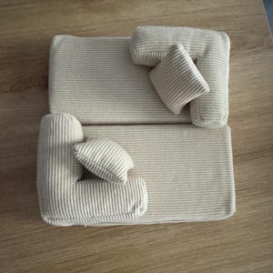 May include: A miniature beige corduroy sofa with two L-shaped sections and two small square pillows. The sofa is placed on a wooden surface. The corduroy fabric has a ribbed texture.