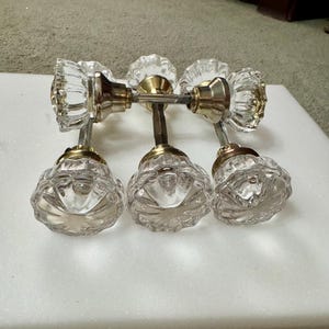 May include: Six clear glass door knobs with brass and silver-toned hardware. The knobs have a faceted, floral-like design. The hardware includes the spindle and mounting plates. The knobs are arranged on a white surface.