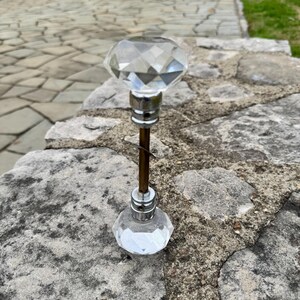 May include: A clear, faceted crystal door knob with a silver base and a bronze rod. The knob is diamond-shaped and reflects light. The image is taken outdoors.