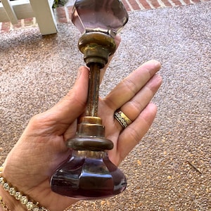 May include: A vintage door knob set featuring two faceted, translucent purple glass knobs connected by a central metal spindle. Each glass knob is secured to the spindle with a tarnished brass-toned metal collar, showing signs of age and wear.