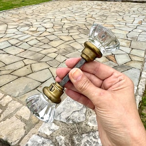 May include: A clear glass and brass door knob with a faceted design. The knob has a brass base and a threaded rod. The background shows a stone walkway and steps.
