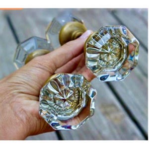 May include: Clear glass door knobs with brass hardware. The knobs have an octagonal shape with a starburst design in the center. The glass is faceted, creating a sparkling effect. The knobs are held in a hand.