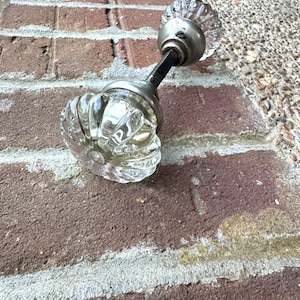 May include: A vintage door knob with clear, ribbed glass knobs and a brushed metal base. The door knob is attached to a black metal rod. The glass knobs have a flower-like design. The door knob is resting on a red brick surface.