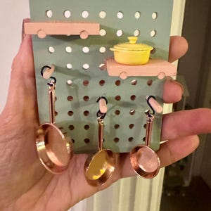 May include: A miniature kitchen set featuring a light green pegboard with small copper-colored pans and a yellow pot. The set includes two wooden shelves and three hanging pans, creating a charming, decorative display.