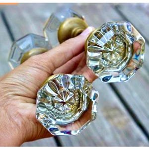 Könnte beinhalten: Klare, facettierte Türknöpfe aus Glas mit messingfarbenen Basen. Die Knöpfe haben eine achteckige Form und reflektieren das Licht, wodurch ein funkelnder Effekt entsteht. Mehrere Knöpfe sind zu sehen.