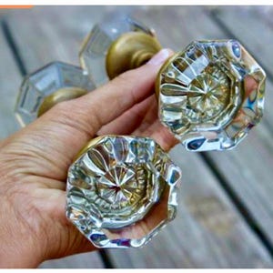 May include: Two octagonal glass door knobs featuring a clear, faceted surface that reflects light. Each knob is attached to a circular brass-toned metal base, showcasing a classic design with a transparent, multi-sided glass handle structure.