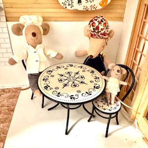 May include: Three plush mouse dolls sitting at a round metal table with a white top featuring black floral patterns. One mouse wears a white chef hat and jacket, another wears a floral headscarf and dark apron, and the smallest wears a white dress.
