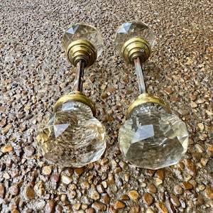 May include: Two clear, multi-faceted glass door knobs featuring a geometric, diamond-like cut. Each knob is attached to a brass-toned metal base and a central spindle, showcasing a reflective, transparent surface with metallic accents.