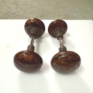 Puede incluir: Dos pomos de puerta antiguos con un diseño marrón veteado. Los pomos de forma ovalada están unidos a una varilla metálica. La ferretería tiene una estética vintage.