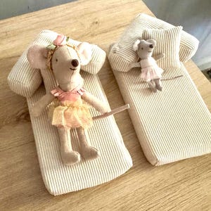 May include: Two plush mouse dolls on corduroy beds. One mouse wears a pink and yellow dress with a floral headband. The other mouse wears a pink dress. The beds have matching pillows and are on a wooden surface.