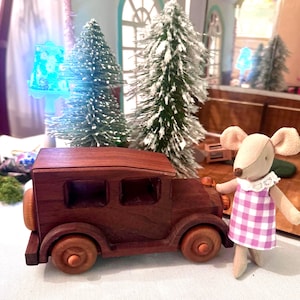 May include: A dark brown wooden toy car with four wheels and a spare tire on the back, positioned next to a plush mouse figurine wearing a white-collared dress with a purple and white checkered pattern, standing in front of two small snowy pine trees.